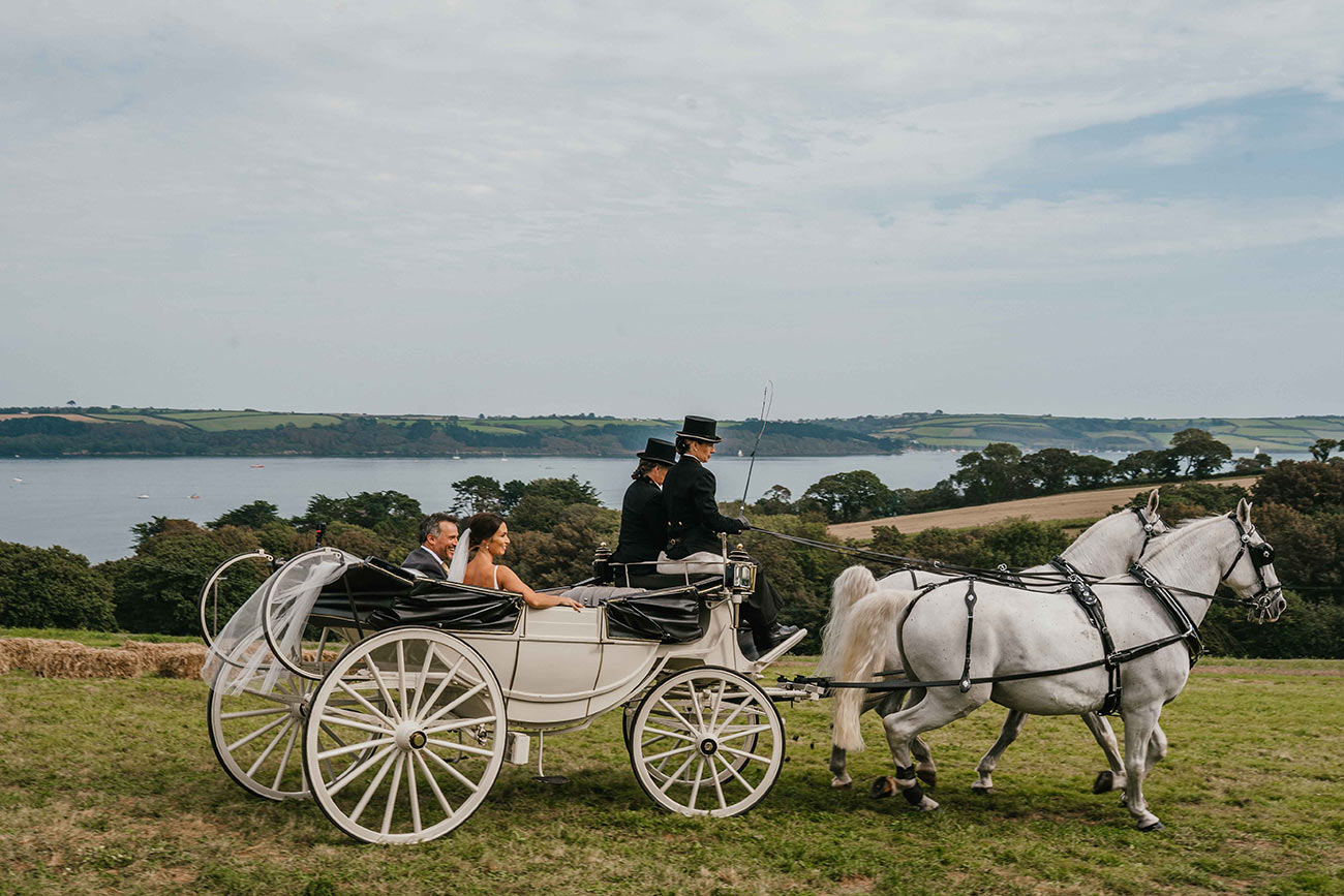 Bride and groom ride in a horse and carriage pulled by two white horses with a stunning view of the sea in the background