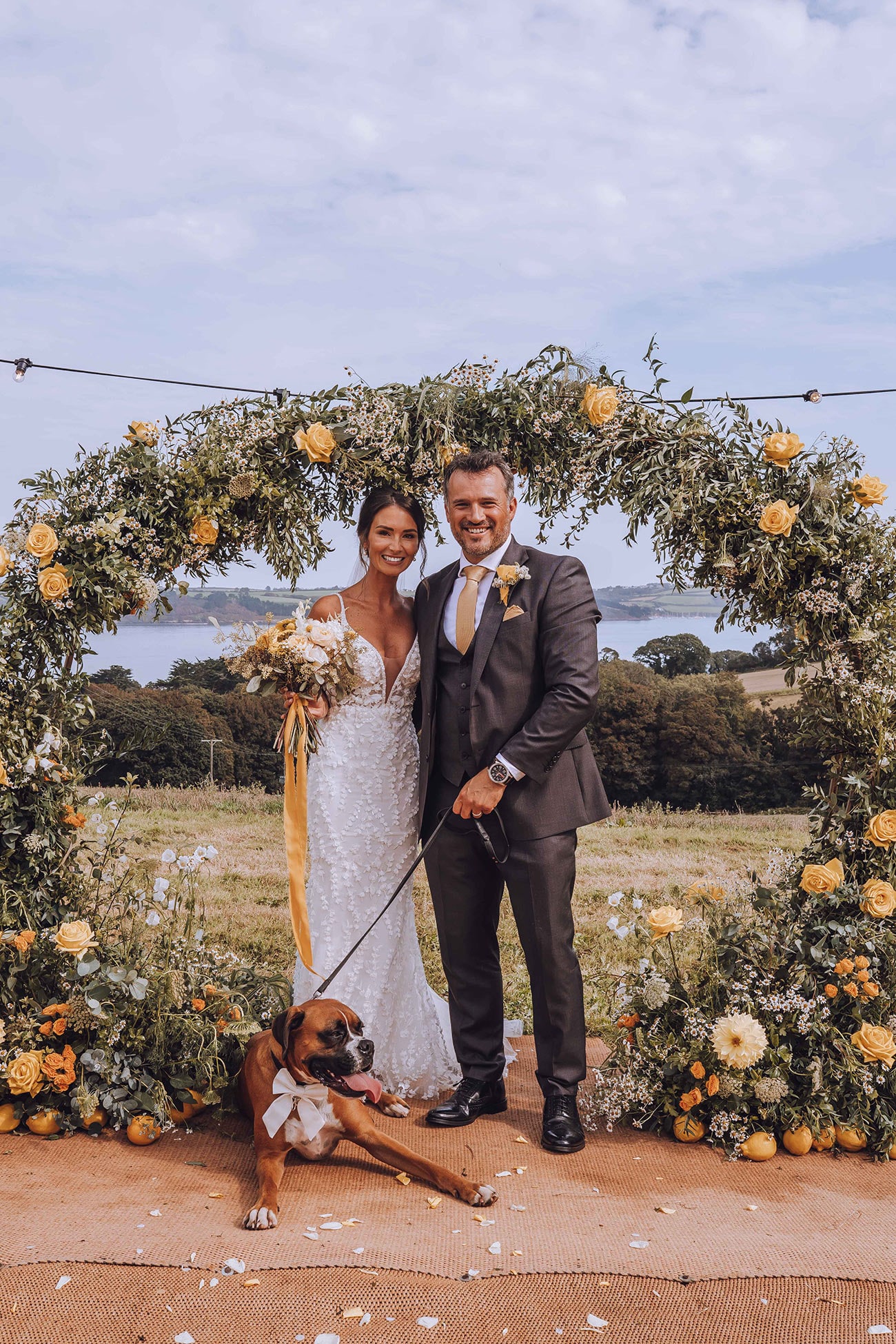Bride and groom stand pose under a yellow and white floral arch with their dog