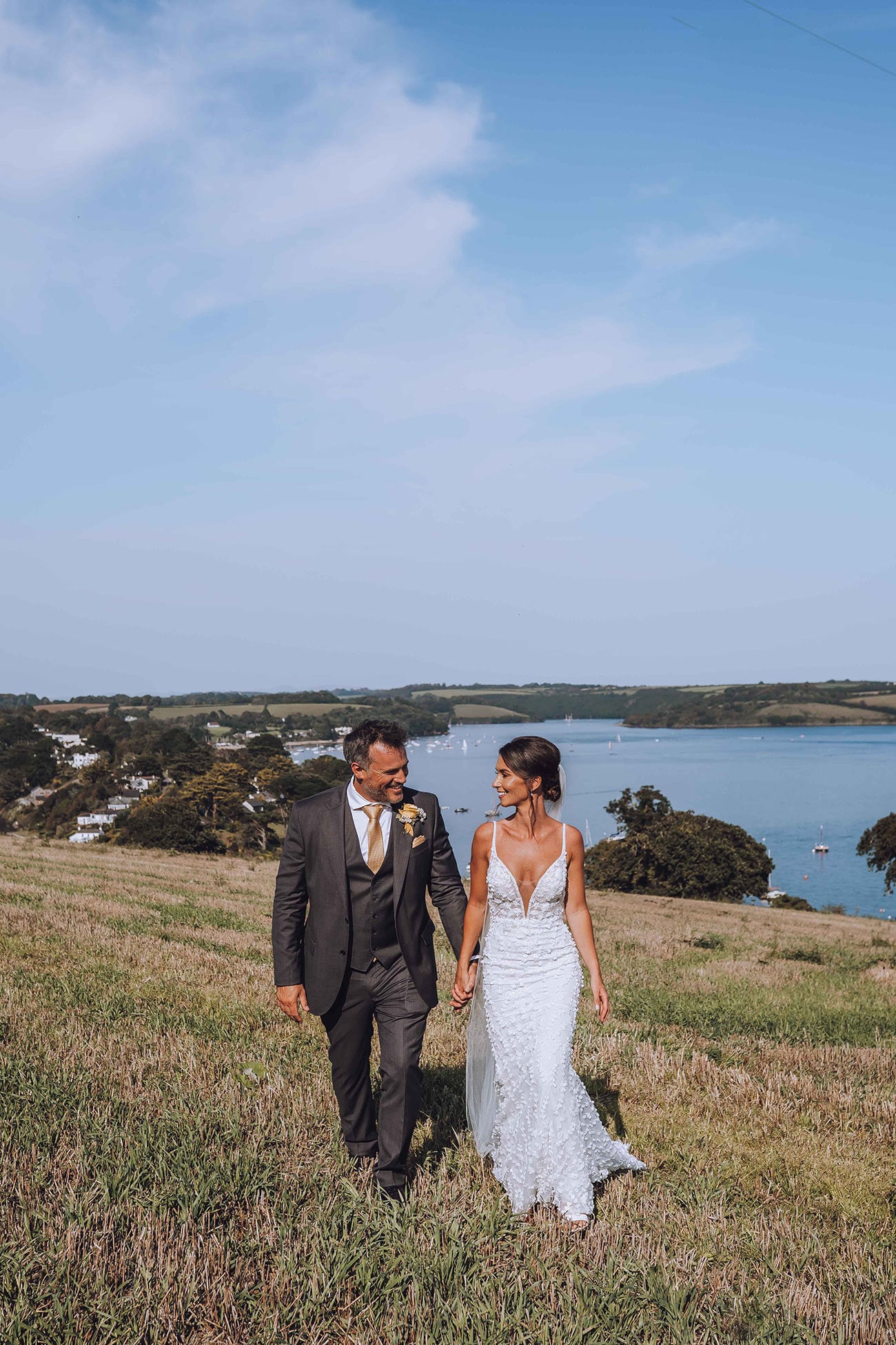 The bride and groom smiling, at each other, walk hand in hand across the field with a coastal backdrop behind 