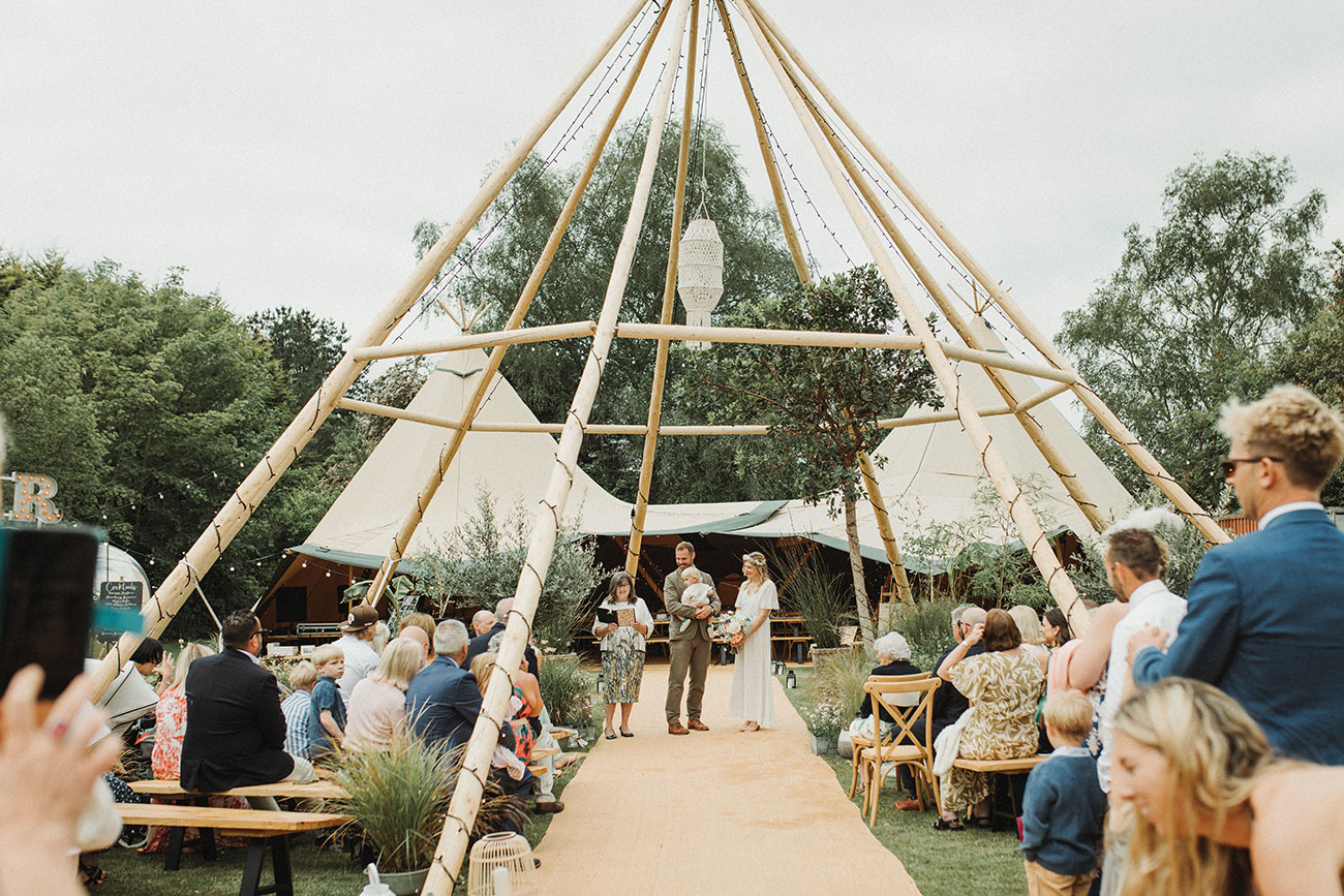 Outdoor wedding ceremony in front of tipi with guests sitting on wooden benches