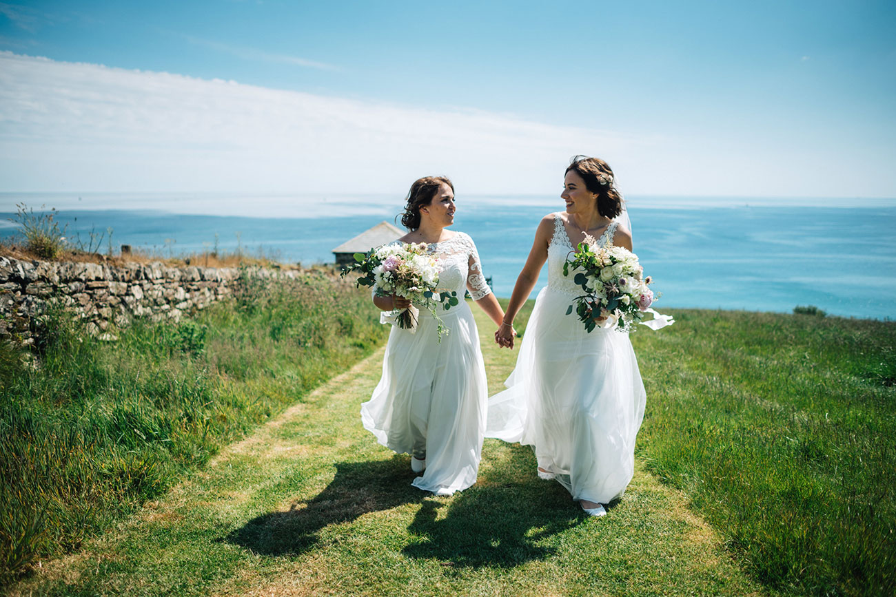 Two brides holding hands and wedding bouquets walking with a sea view behind them