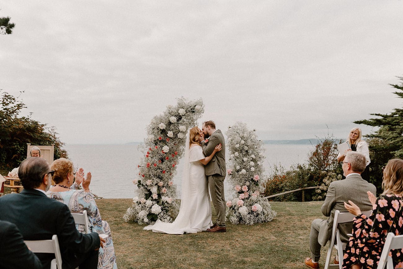 Bride and groom having their first kiss as a married couple under a floral arch in front of their clapping guests
