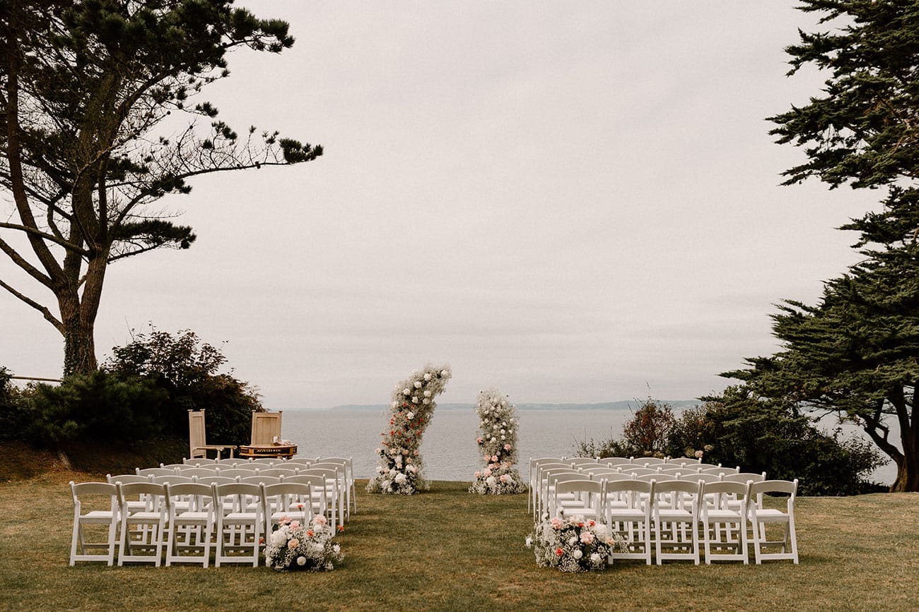 Outside wedding ceremony set up with white wooden chairs and floral arch