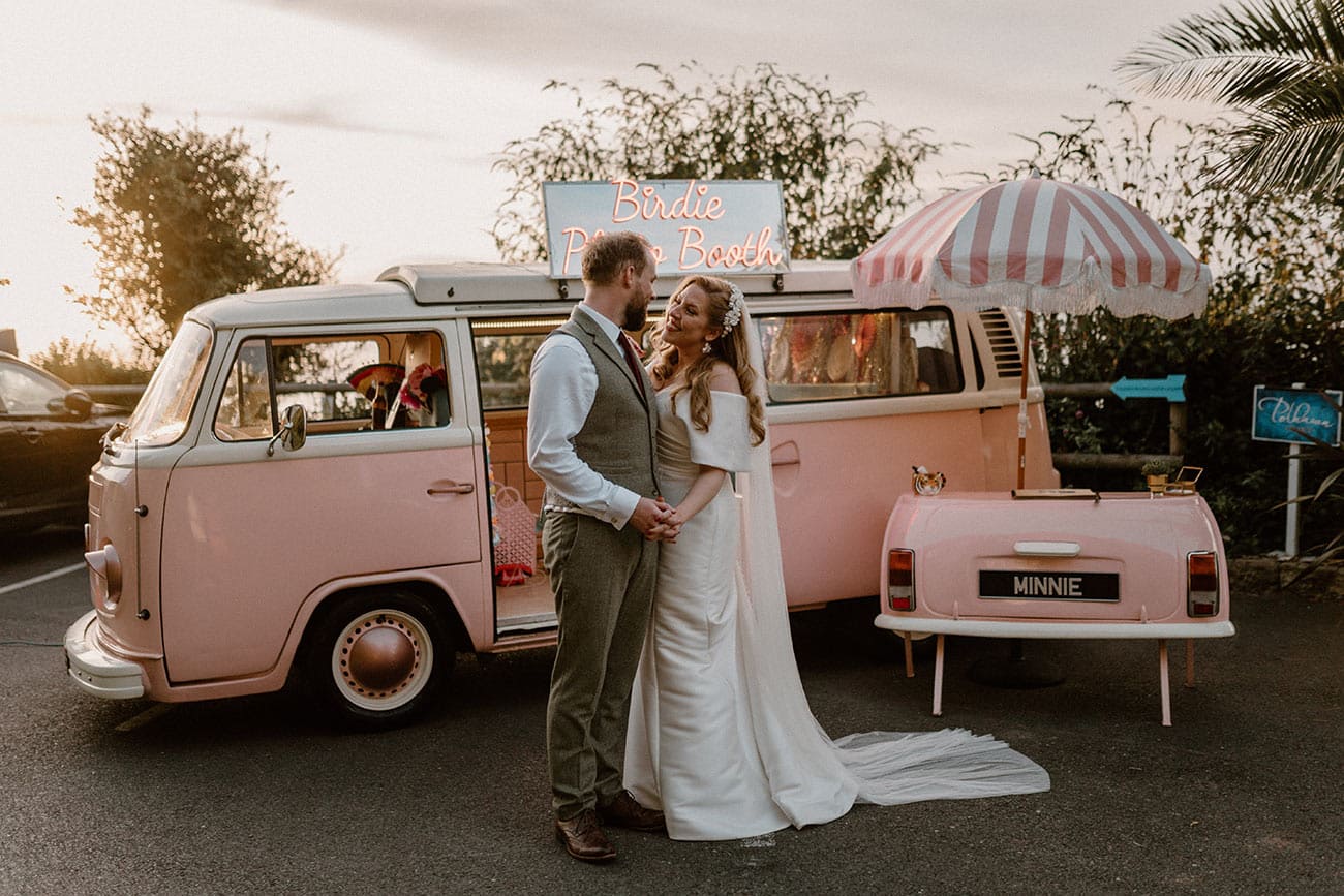 Bride and groom posing in front of a pink, retro Photo Booth camper van