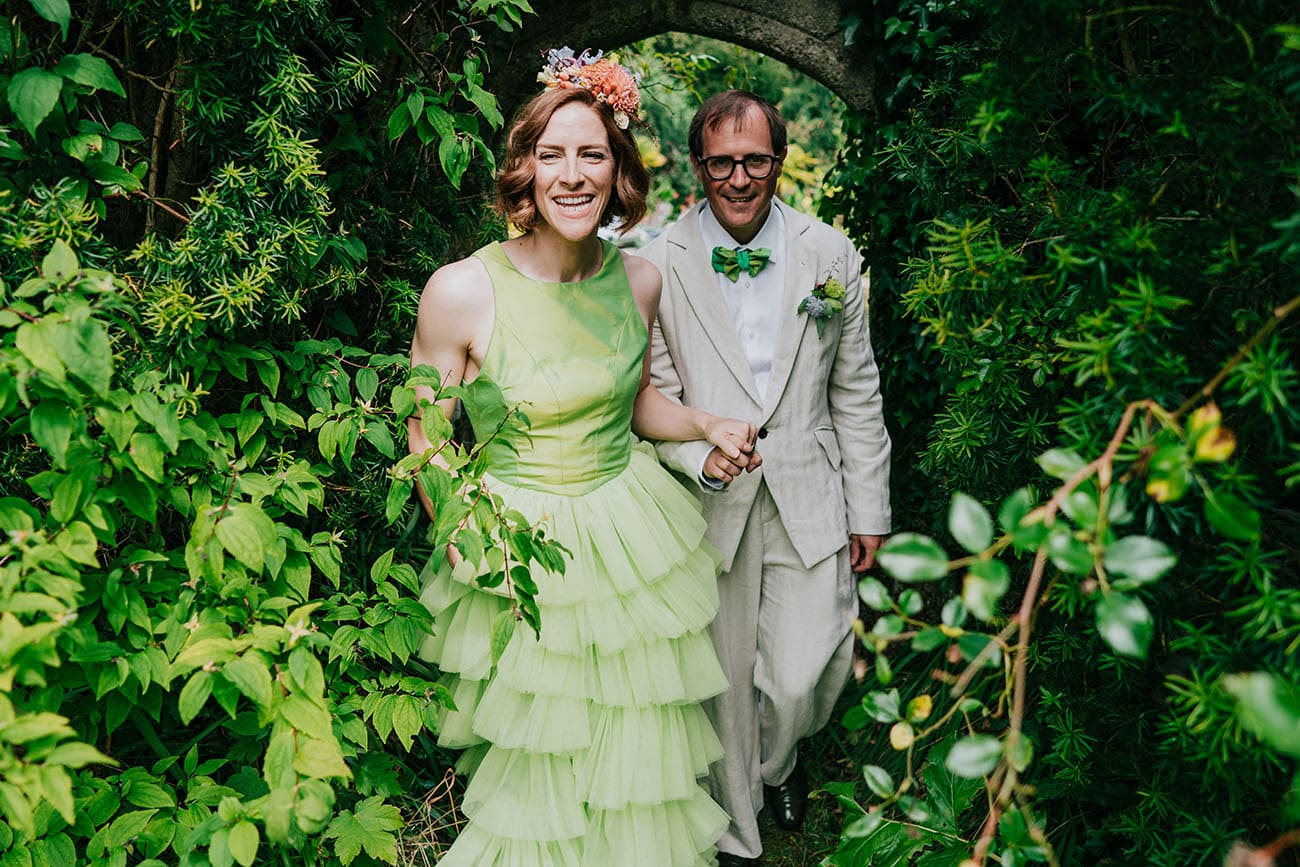Bride in colourful green bridal gown and groom walk hand in hand under an arch of foliage